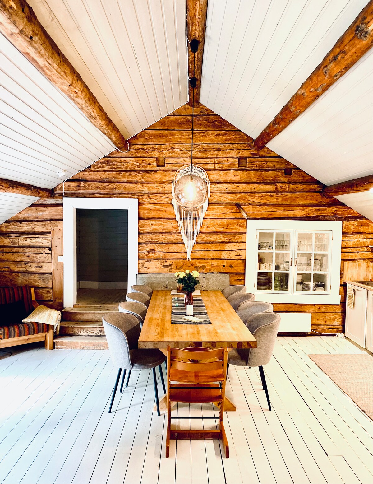 A warm dining area features a large wooden table surrounded by eight upholstered chairs. The ceiling showcases wooden beams and a distinctive pendant light, while a rustic cabinet is visible in the background. The floor is light-colored, enhancing the inviting atmosphere.