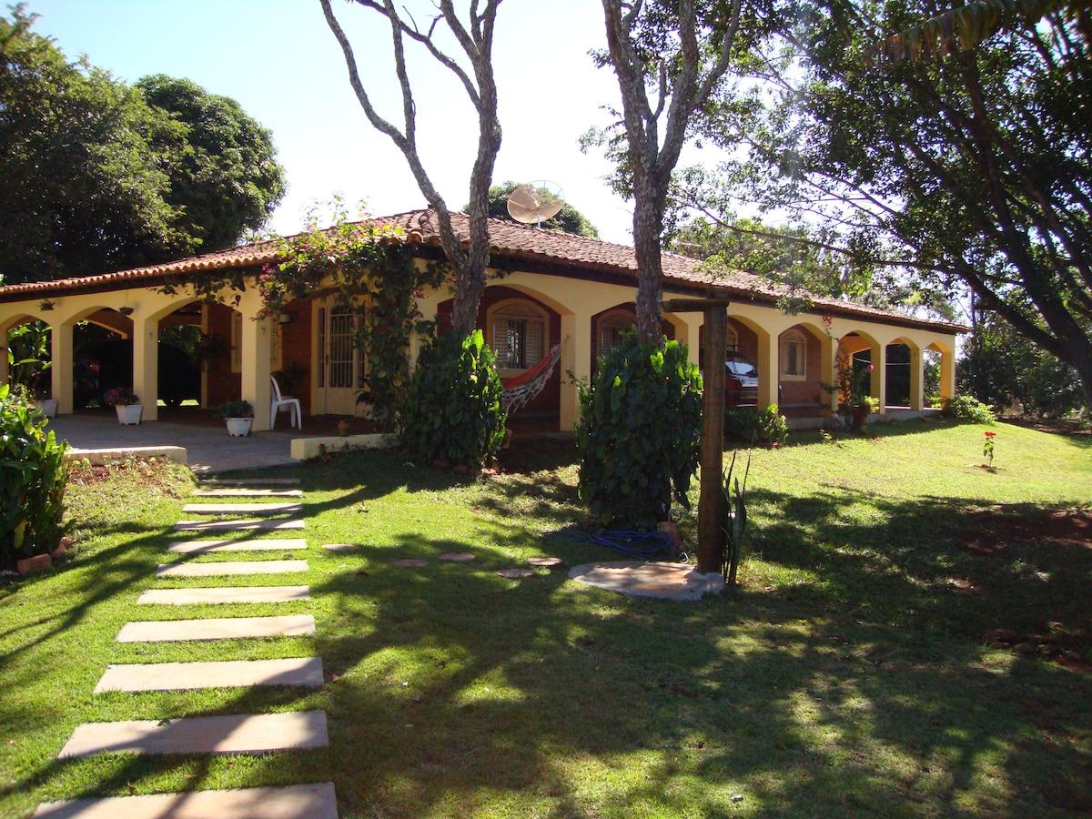 A single-story house is surrounded by green grass and mature trees. A pathway of stepping stones leads to the entrance, where a covered porch can be seen. The structure features a tiled roof and large windows, contributing to a spacious ambiance.