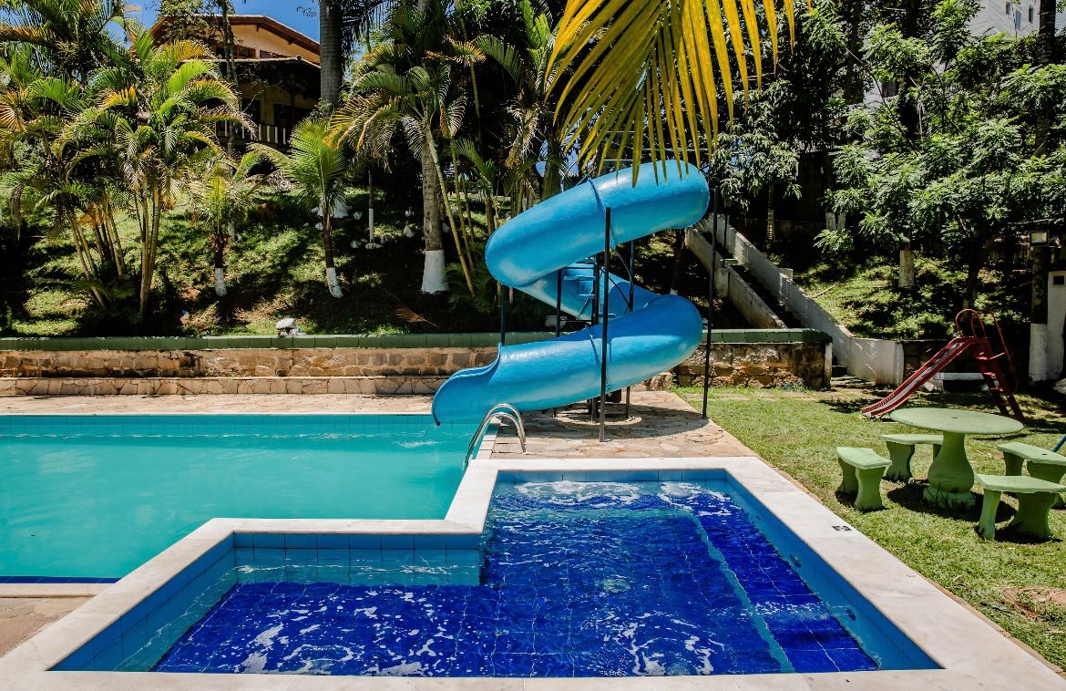 A large swimming pool is complemented by a blue waterslide, surrounded by lush greenery. A smaller hot tub is visible to the side, featuring blue tiles. Nearby, a playground is indicated with colorful equipment, providing additional recreational options.