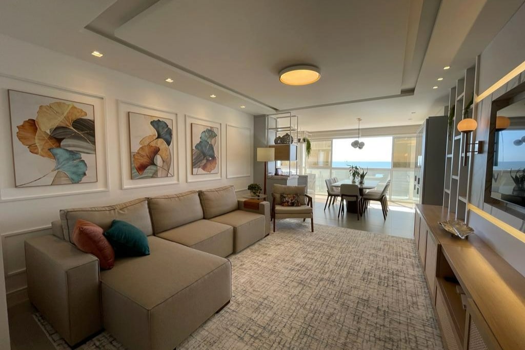 A modern living area features a spacious sectional sofa with decorative pillows, complemented by artwork on the walls. A dining table and chairs are visible in the background, with large windows allowing natural light and providing a view of the ocean.