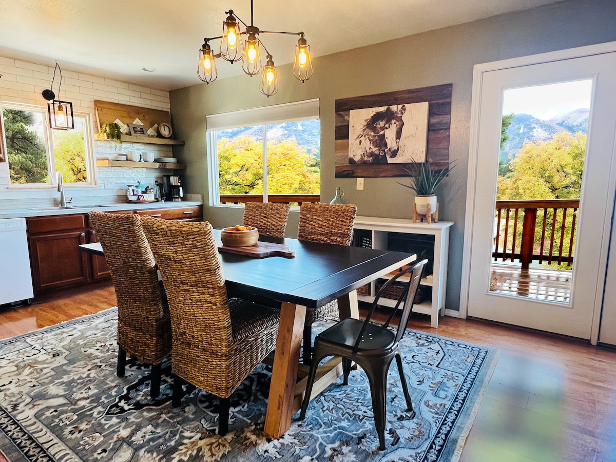 A dining area features a wooden table surrounded by six woven chairs, complemented by a woven rug. Large windows invite natural light while offering views of the landscape. A sideboard holds decorative items, and a set of pendant lights hangs above the table.