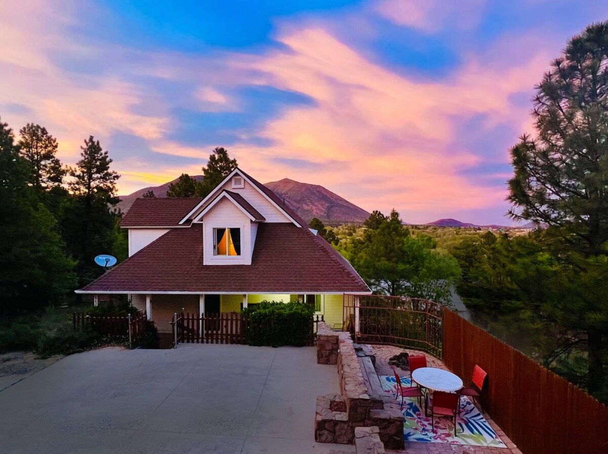 A charming house is set against a colorful sky during sunset, surrounded by tall pine trees. The exterior features a sloped roof and a welcoming front porch. A patio area with a table and chairs is visible, along with a decorative stone wall.
