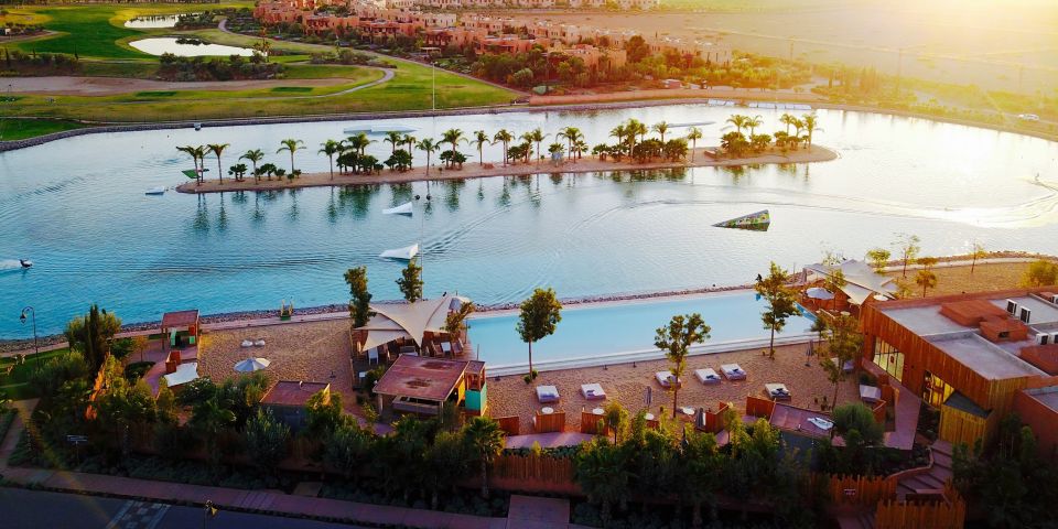 An aerial view captures a serene landscape featuring a winding waterway bordered by sandy areas and palm trees. Sun loungers are arranged along the edges, with vibrant greenery visible in the background, while water sports equipment is situated nearby.
