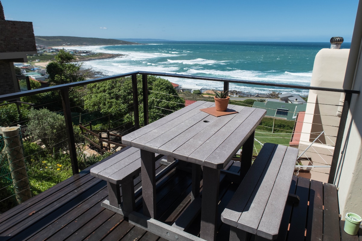 A wooden deck features a dining table and benches, offering a space for outdoor seating. The expansive view of the ocean reveals rolling waves and a distant coastline. Lush greenery is present in the foreground, enhancing the serene atmosphere of the area.