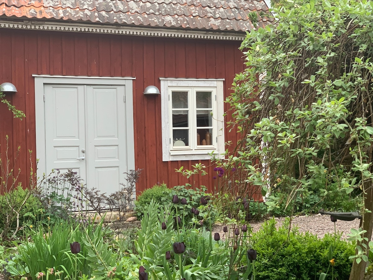 A traditional red wooden guest cabin is framed by a lush garden, featuring various plants and flowers in rich green hues. Two white double doors provide an inviting entrance, complemented by a window that adds charm. Soft natural light highlights the serene surroundings.
