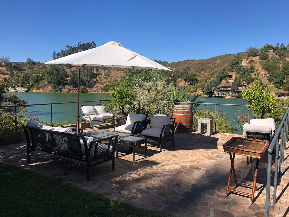 A spacious terrace overlooks the lake, featuring comfortable seating under a large umbrella. A stone pathway is surrounded by greenery and a few decorative plants. A small table and barrel add rustic charm to the outdoor space.