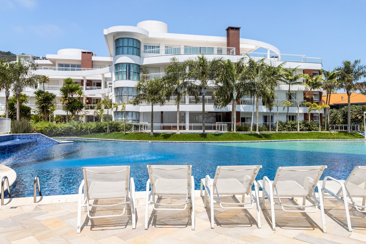 A luxurious resort is displayed, featuring a spacious swimming pool surrounded by well-maintained palm trees. Several lounge chairs are positioned near the water's edge, providing a relaxing spot to enjoy sunny days. Clean lines and modern architecture characterize the multi-level building in the background.