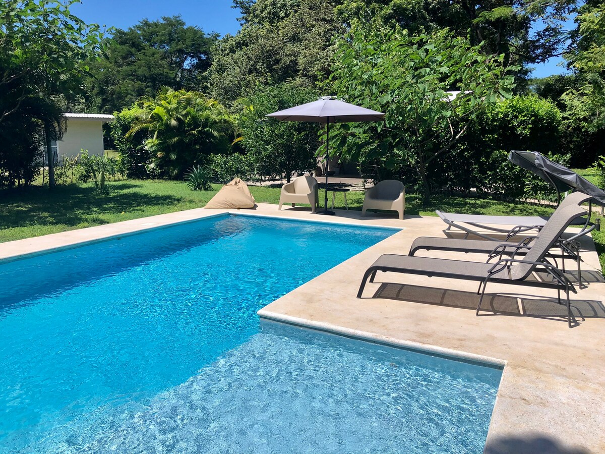 A refreshing swimming pool is surrounded by a well-maintained garden, featuring several lounge chairs and a shaded table with an umbrella. Nearby, a day bed provides a relaxing spot within the lush greenery.