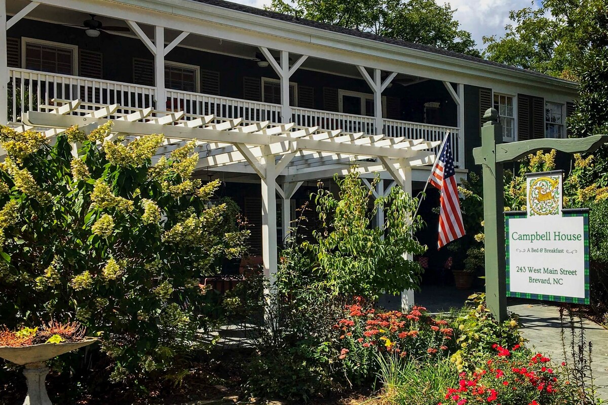 The exterior of Campbell House is showcased, featuring a spacious covered porch supported by white pillars. Lush greenery and colorful flower beds surround the entrance, while an American flag hangs proudly. A sign indicating the location adds a welcoming touch.