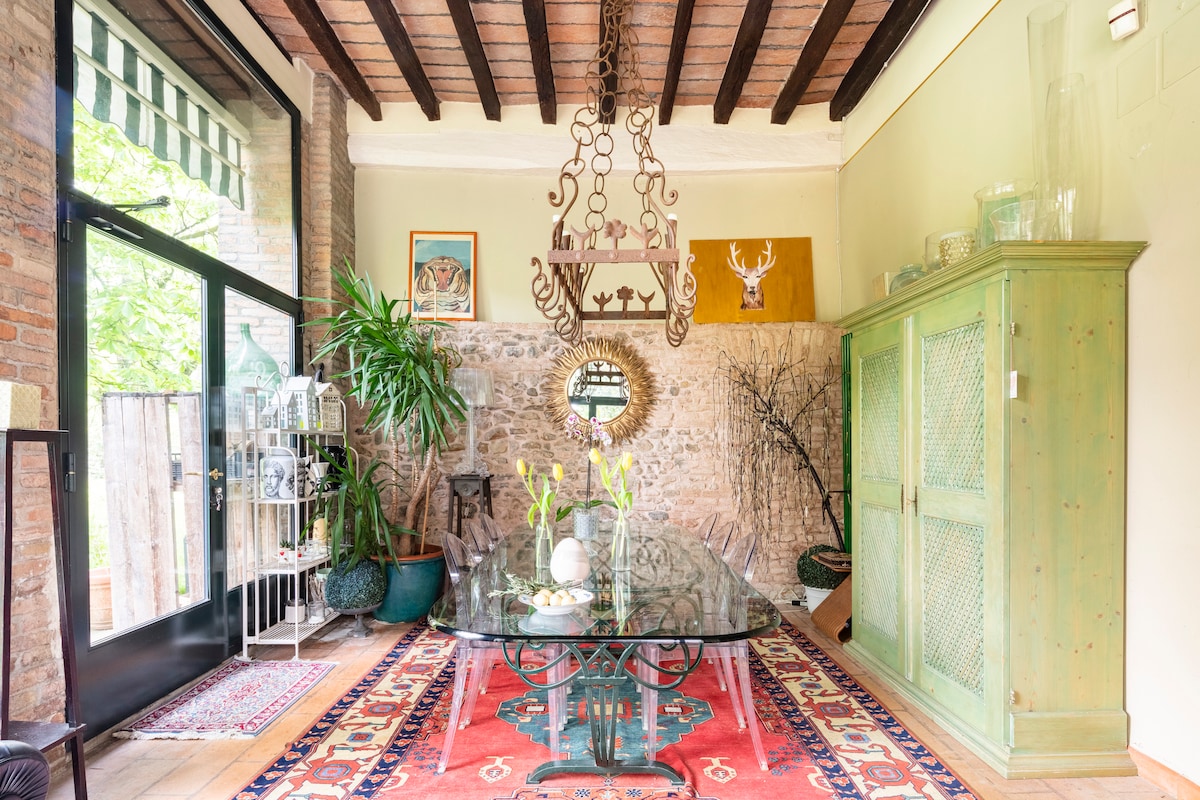 A bright dining area features a large glass table surrounded by wooden chairs. A decorative chandelier hangs from the ceiling with exposed wooden beams. A vibrant area rug adds warmth to the space, complemented by greenery from potted plants and artworks on the walls.