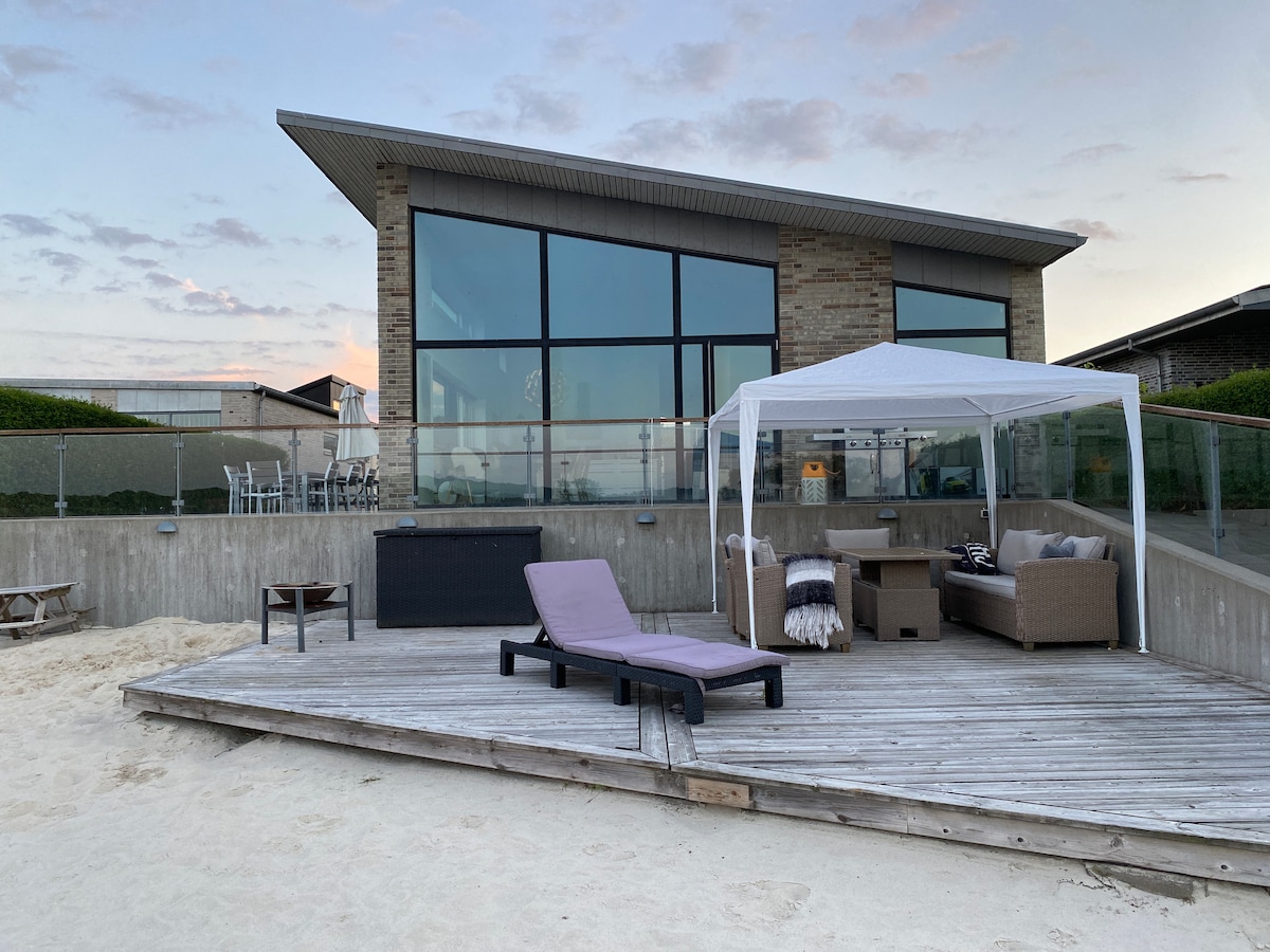 A modern villa is showcased, featuring large glass windows and a covered seating area on a wooden deck. A lounge chair is positioned for relaxation by the beach, with soft sand surrounding the space. Outdoor furniture invites gatherings while the sky reflects soft evening hues.
