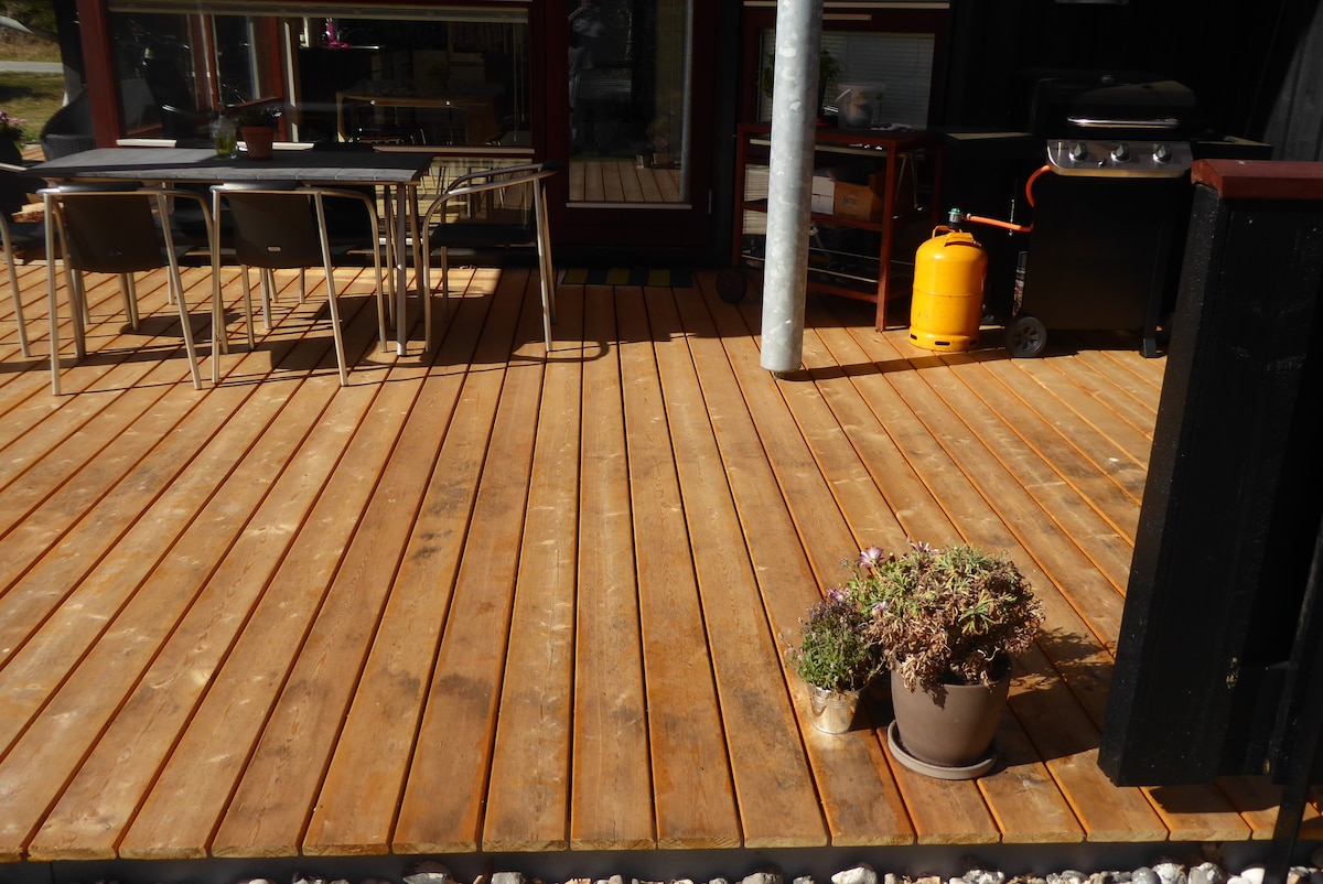 A wooden deck is shown, featuring a table and several chairs. A gas grill is positioned alongside a propane tank. A potted plant with flowers adds a touch of greenery to the space. The deck area is well-lit, indicating a welcoming outdoor setting.