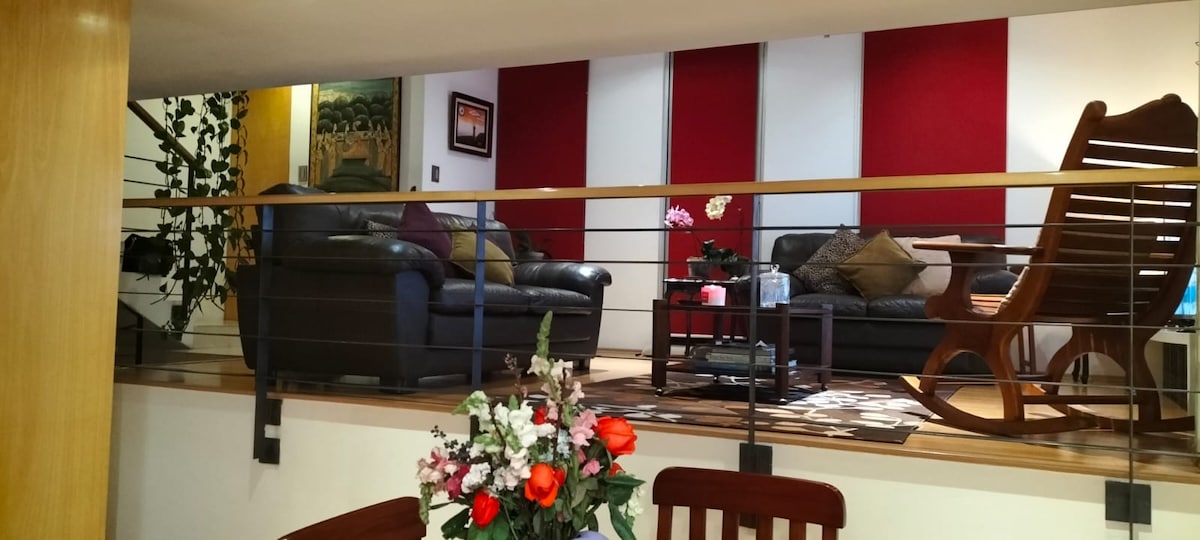 A welcoming living area is highlighted with dark leather sofas and a wooden rocking chair. A coffee table sits at the center, surrounded by a decorative rug. Red accent walls create a vibrant backdrop, complemented by indoor plants and a floral arrangement on a dining table.