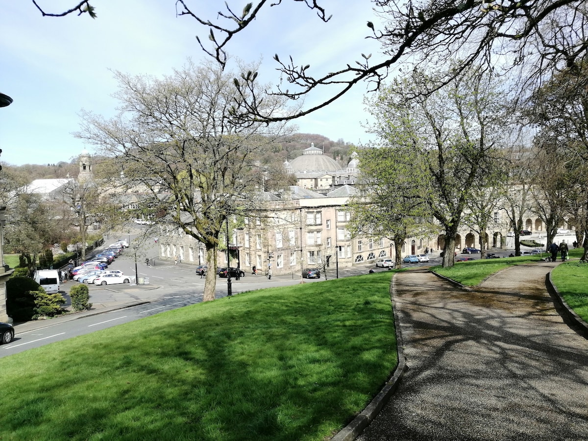 The image captures a scenic view of a park area with well-maintained green lawns and winding pathways. Surrounding buildings with classic architecture are visible in the background, along with trees that provide shade. A glimpse of distant hills adds depth to the landscape.