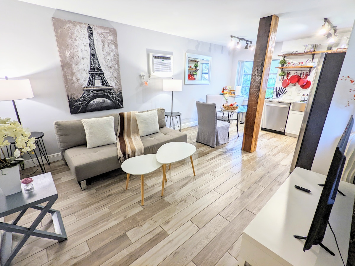 The living area is furnished with a modern sofa bed and a pair of minimalist tables. A large artwork of the Eiffel Tower adorns the wall, while natural light filters through nearby windows. The open layout connects to the kitchen, which features stainless steel appliances and bar stools.