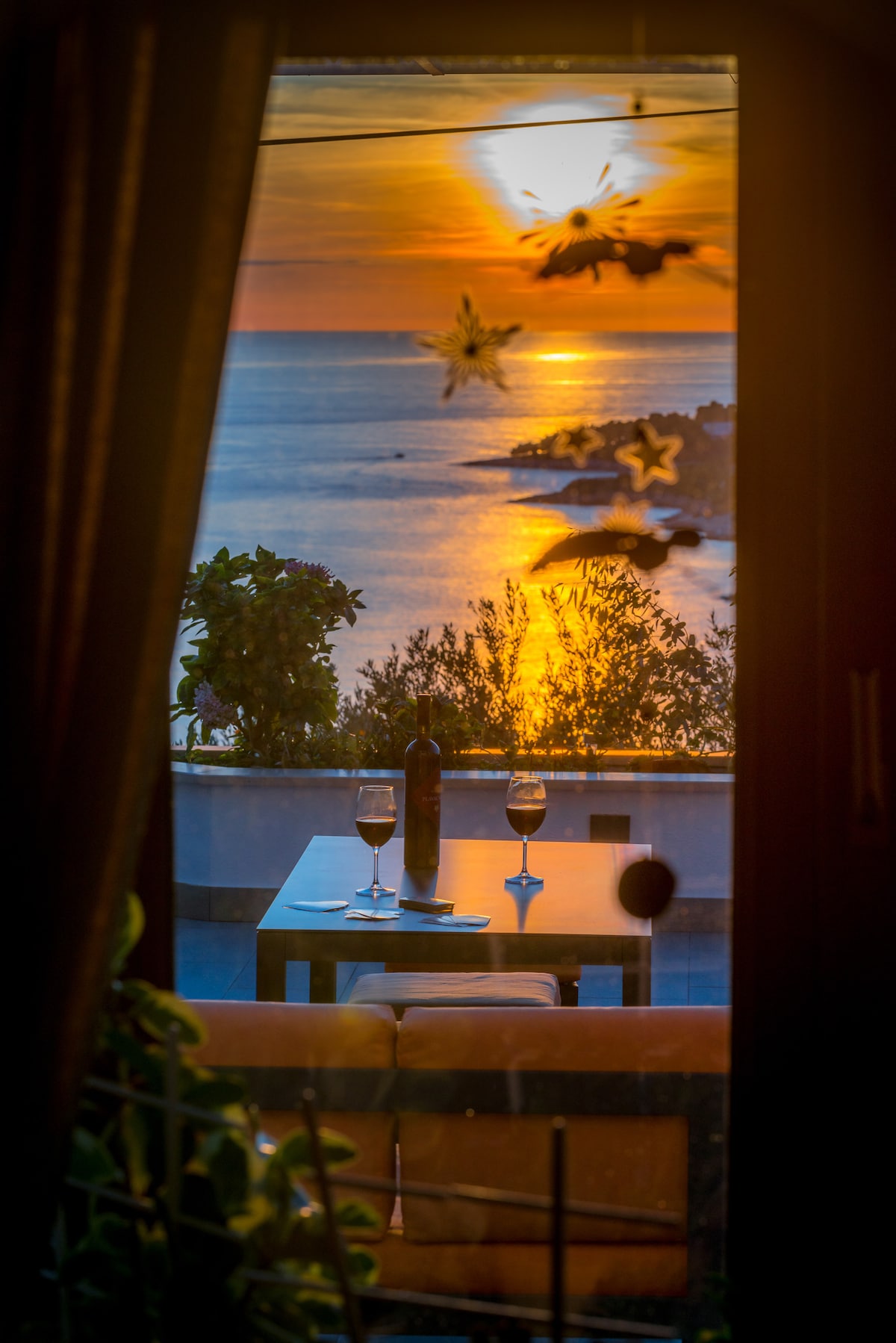 A serene outdoor dining area is visible through large glass doors, featuring a table set for two with wine glasses. The sunset casts warm tones over the sea, enhancing the beautiful view of the coastline.