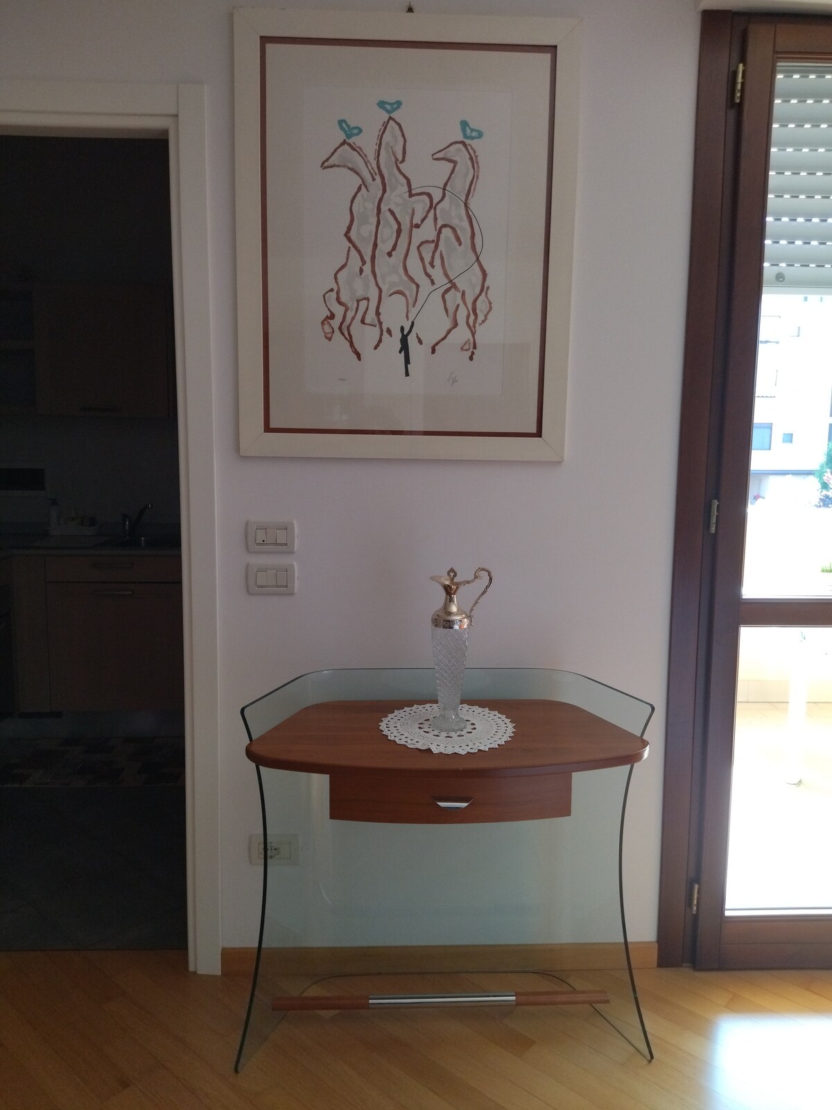 A small, glass-top table with a wooden drawer is displayed against a light-colored wall. A decorative vase sits on the table, accompanied by a delicate lace doily. Artwork featuring abstract figures hangs above, complementing the contemporary design.