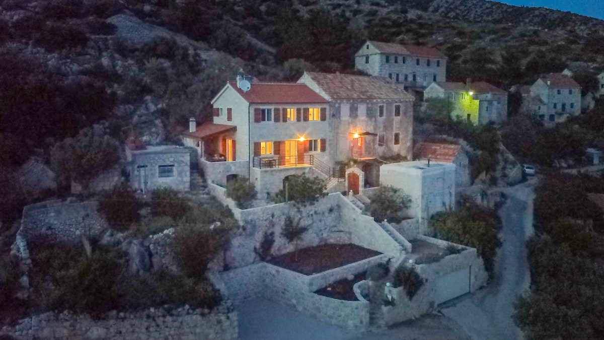The house is nestled amidst a hillside village, showcasing a blend of stone and traditional architecture. Soft lights illuminate the exterior, highlighting the warm welcoming entrance. Surrounding greenery and stone pathways contribute to the home’s rustic charm, set against a backdrop of rolling hills.