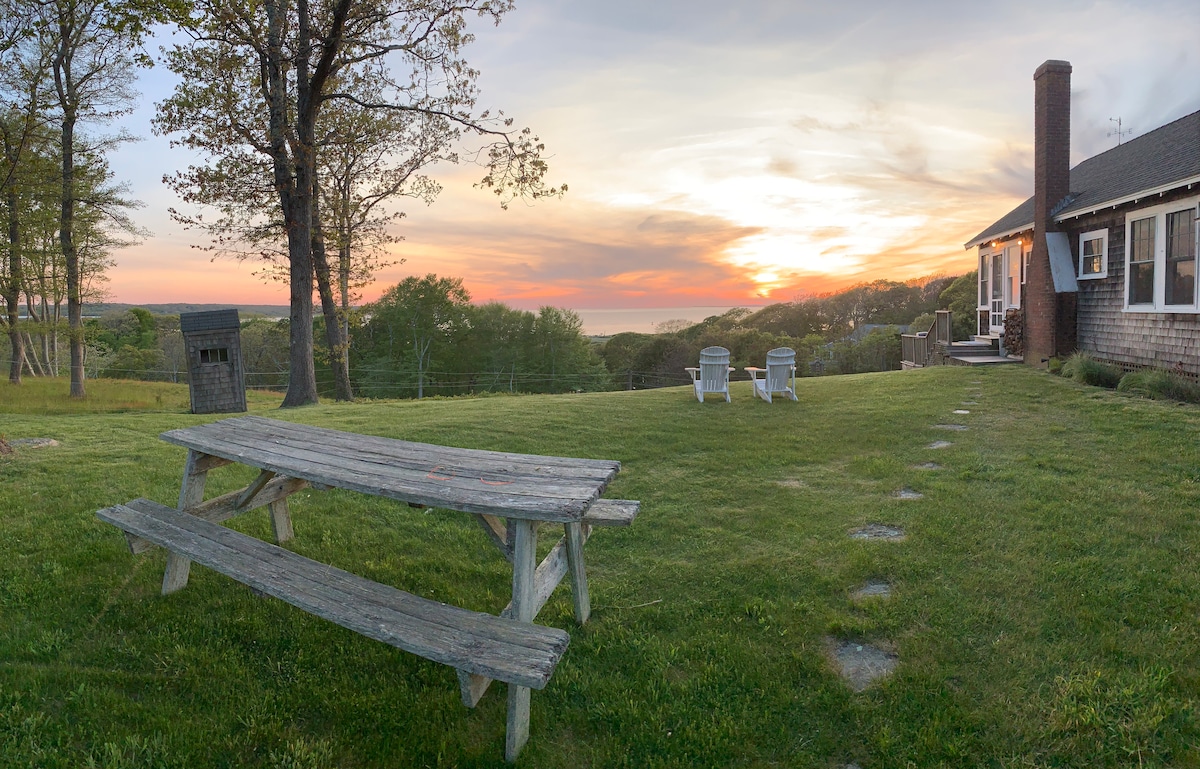 An outdoor scene showcases a picnic table made of weathered wood, surrounded by a grassy area. Two white adirondack chairs are positioned nearby, facing a breathtaking sunset over the horizon. The rustic cottage is visible in the background, along with trees that frame the view.