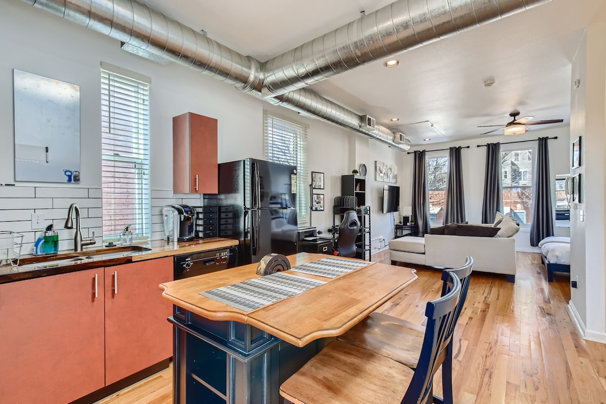 An open living space features a kitchen with modern appliances, including a black refrigerator and a microwave. A wooden dining table is positioned nearby, while natural light enters through large windows. A comfortable seating area with a neutral sofa is visible in the background.