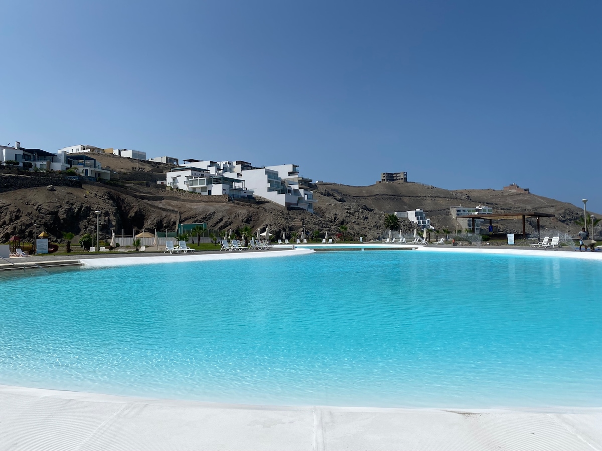 A large, inviting pool reflects the clear blue sky, surrounded by manicured greenery and sun beds. The tranquil setting features modern houses atop a hillside in the background, enhancing the peaceful atmosphere of the private club.