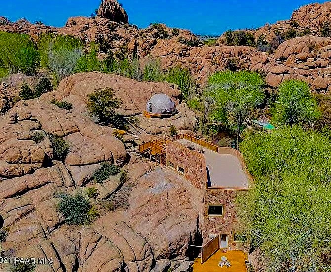 Aerial view of Cave Castle surrounded by large granite boulders and lush greenery. The two-story structure showcases a terrace with seating. An additional dome can be seen nearby, along with seasonal greenery and a pool in the background, all set against a clear blue sky.