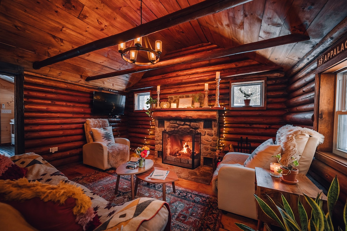Welcome to your cozy cabin retreat. This living room has everything you need to unwind for a perfect night in, from the fireplace and comfortable seating to games, blankets, books, candles, string lights, speaker, and TV. 

PC:@dirtandglass 