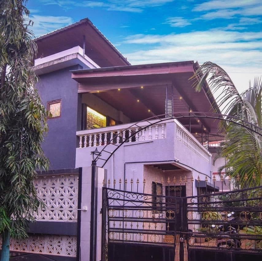 A modern villa is showcased with a two-story structure featuring a mix of dark and light exterior colors. A decorative gate leads to the entrance, while palm trees are visible nearby. The balcony is adorned with subtle lighting, creating an inviting appearance.