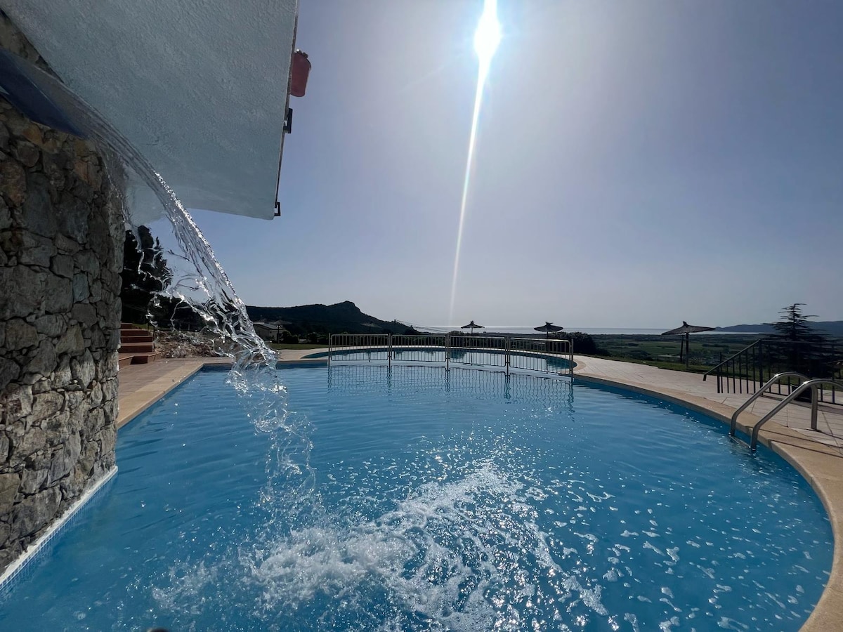 A community swimming pool is visible, featuring clear blue water and a gentle water fountain. Sunlight sparkles on the surface, while lounge chairs are positioned along the edge. Scenic views of the surrounding hills and landscape are in the background, adding a serene ambiance.