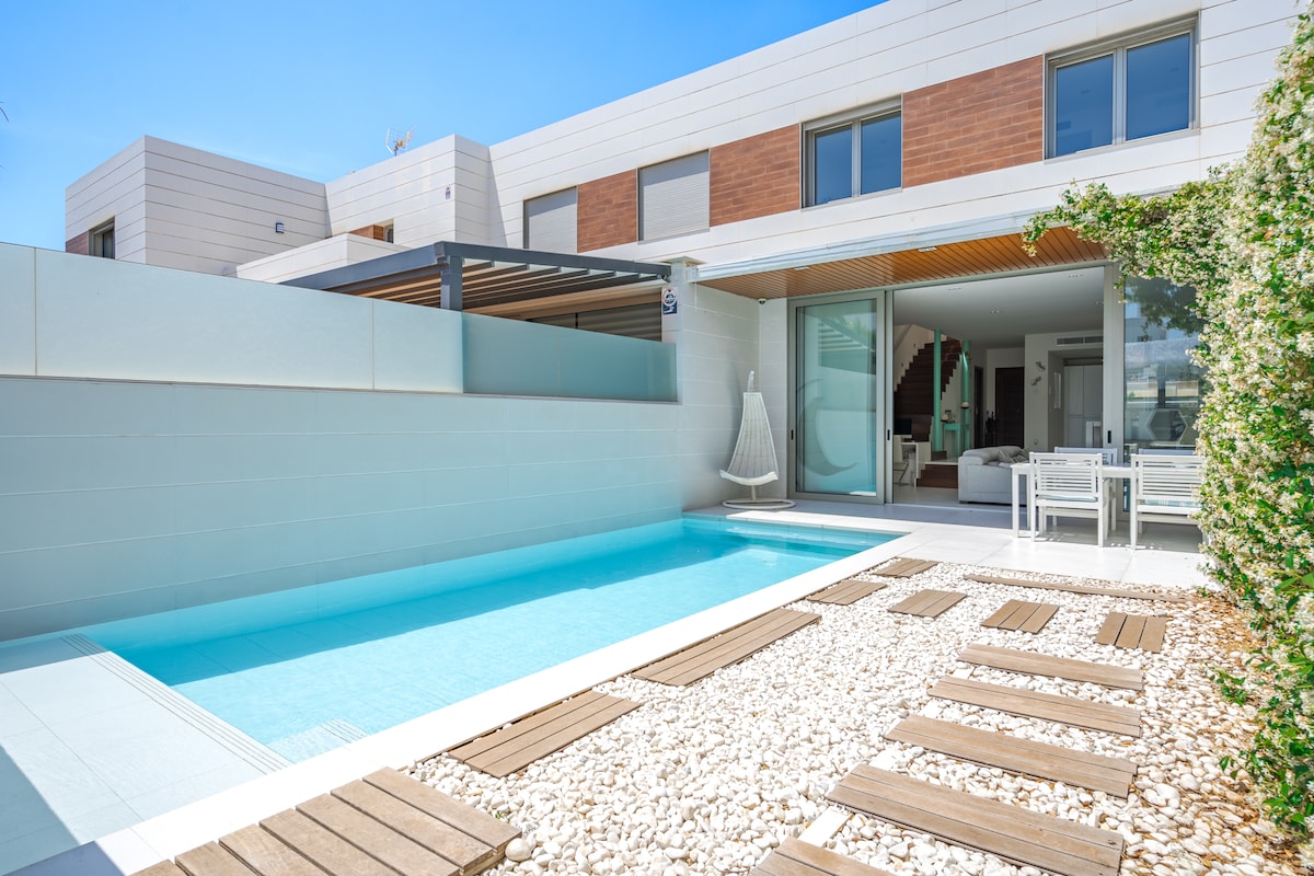 A private pool is framed by smooth stones and wooden decking, creating a serene outdoor space. The house features large glass doors that open to a modern indoor dining area, offering a seamless transition between indoor and outdoor living.
