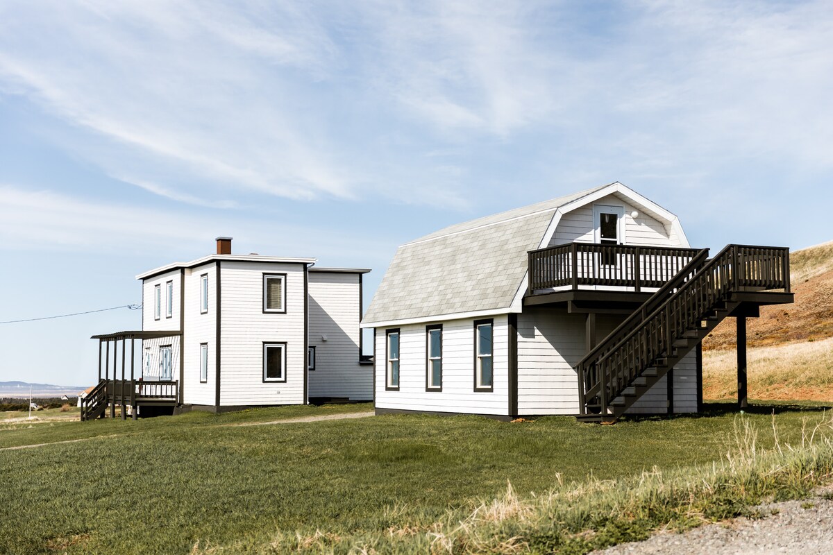 A charming Madelinot home with a white exterior is shown, complemented by a loft above a garage. The two distinct structures are surrounded by a green lawn, set against a backdrop of gentle hills and a clear blue sky.