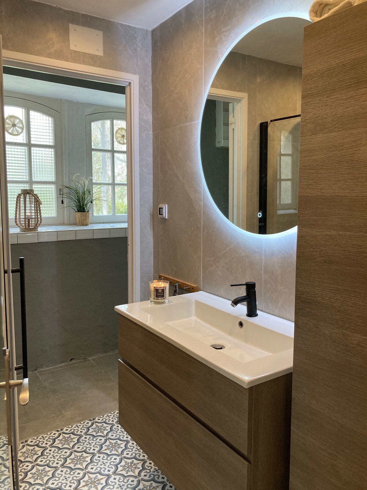 The bathroom features a sleek vanity with a white sink and wooden drawers. A round mirror with ambient lighting is positioned above the sink. Intricate patterned tiles are visible on the floor, while natural light enters through a nearby window.
