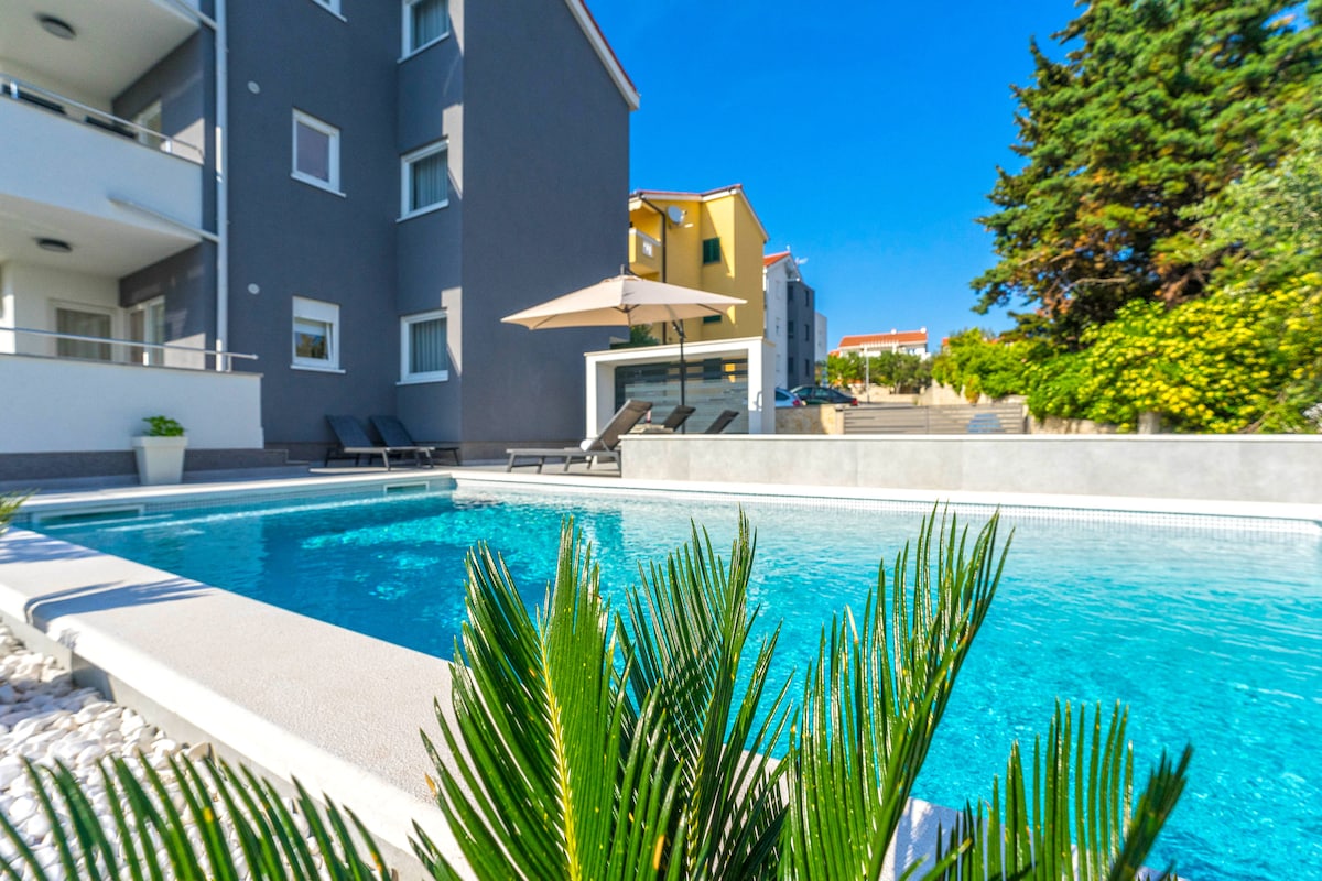 A shared pool area is visible, surrounded by a clean, manicured environment. Sun loungers are positioned alongside the water, and a shaded umbrella provides a relaxing spot. Lush greenery frames the scene, with modern apartment buildings in the background under a clear blue sky.
