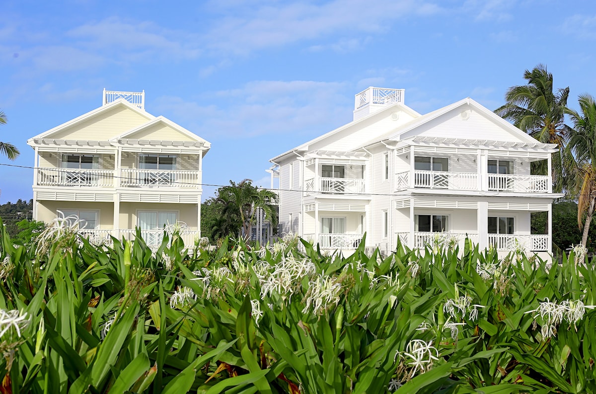 Two identical, modern apartment buildings are set against a blue sky, with palm trees framing the scene. Lush green foliage and white flowers are in the foreground, enhancing the tropical ambiance of the location.