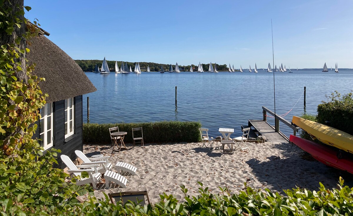 A sandy beach area is framed by a hedge, featuring white lounge chairs and a small table. A cozy beach house is situated nearby, with colorful kayaks stored on the dock. Sailboats can be seen in the calm water under a clear blue sky.