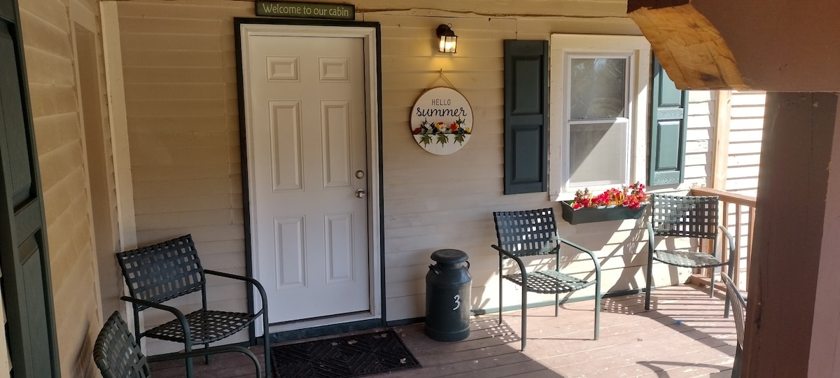 A welcoming porch features a white door with a decorative sign reading 'Hello Summer.' Black seating is arranged around a small table, while potted flowers add color to the surroundings. A trash bin is positioned beside the door, contributing to the neat appearance.