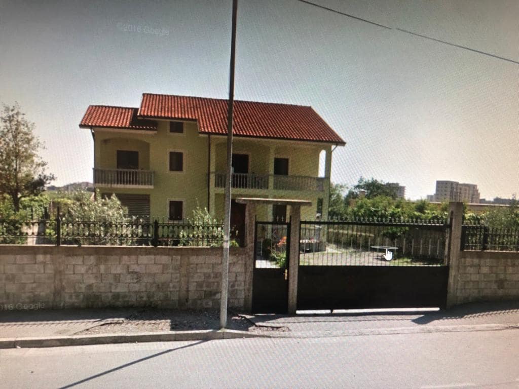A two-story yellow house with a red-tiled roof is surrounded by a low stone wall and fencing. A gated entrance leads to a spacious yard, with greenery visible in the background. The house features a balcony on the upper level, enhancing its inviting exterior.