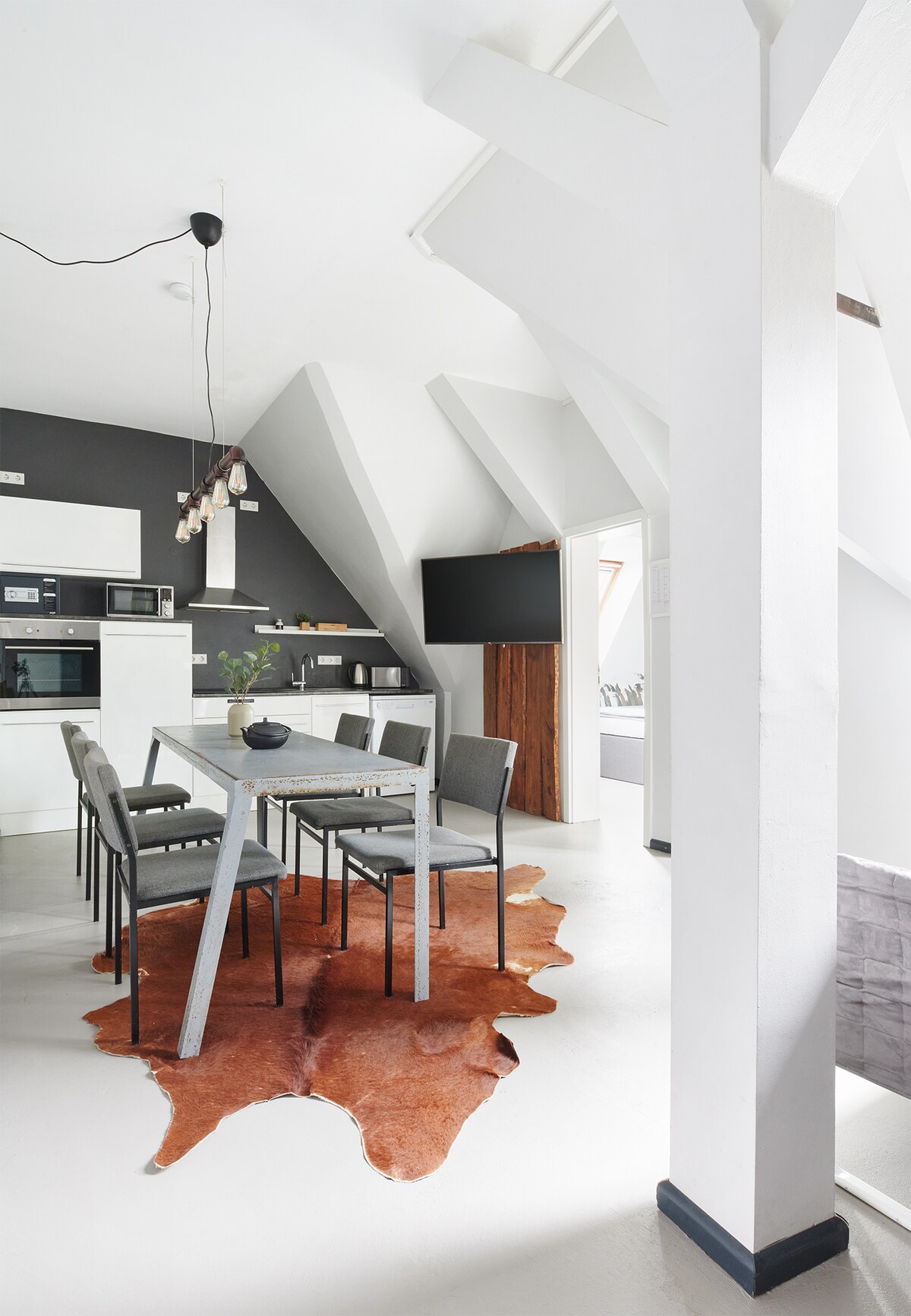A modern kitchen and dining area features a sleek table surrounded by six chairs. A distinctive cowhide rug adds texture beneath the table. The space is illuminated by pendant lighting and is complemented by a black accent wall with a mounted TV. White beams create an airy feel.