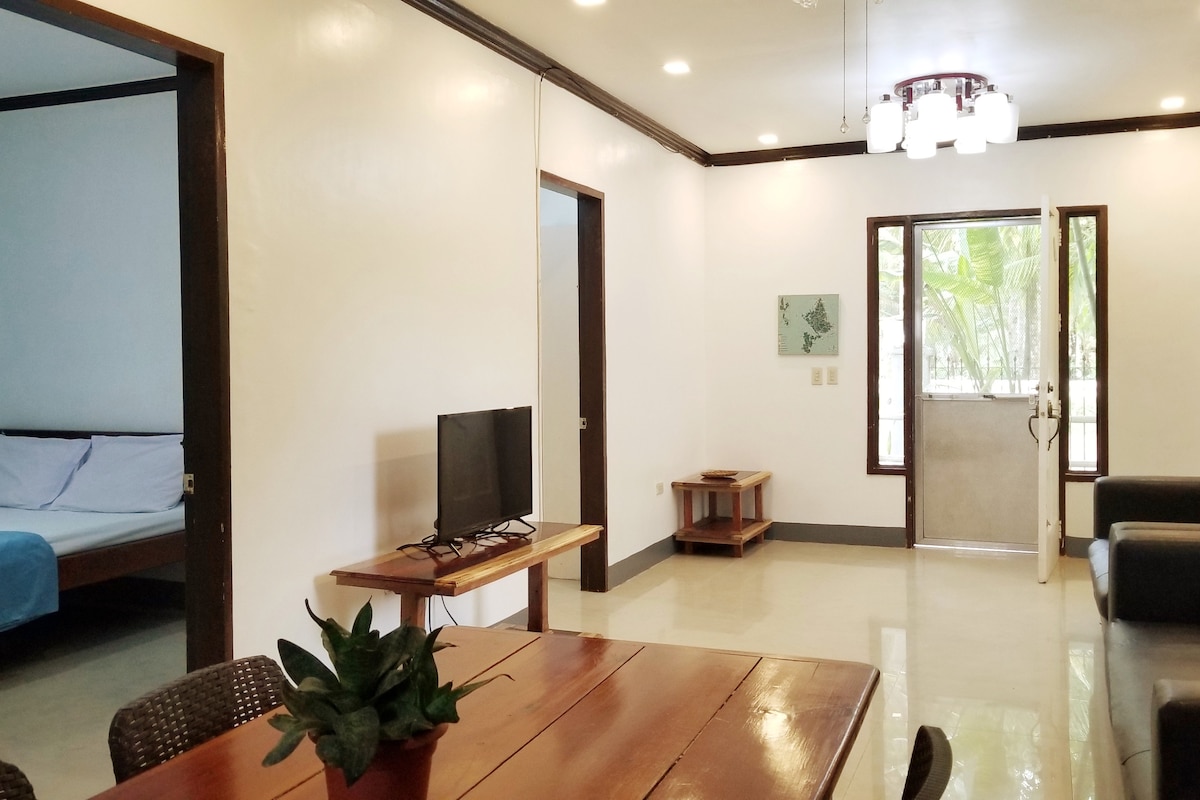 A spacious living area is designed with a wooden dining table and a television. Large windows allow natural light to fill the space, while viewing the garden outside. A comfortable sofa and decorative plant enhance the inviting ambiance. Adjacent, a simple bedroom is visible.