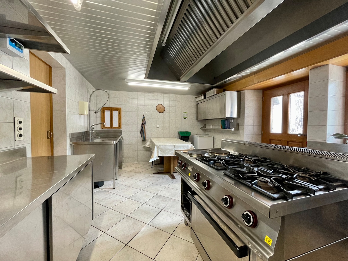 The kitchen is equipped with stainless steel appliances, including a professional stove and a large stainless steel sink. Ample counter space and cabinetry are visible, along with a dining table set for casual meals. Natural light enters through a window, enhancing the workspace.