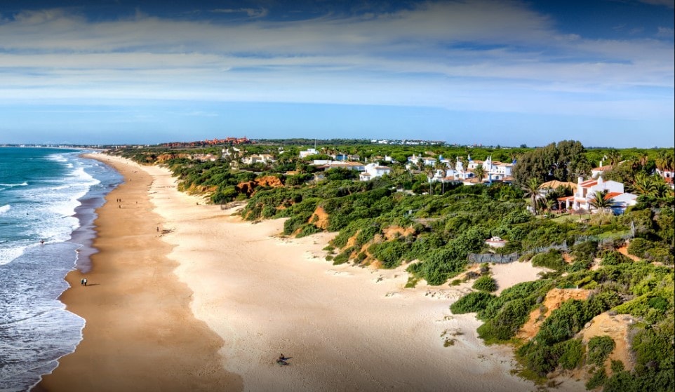 A wide sandy beach stretches along the coastline, bordered by lush greenery. The calm waves gently lap at the shore, with a few figures strolling through the sand. In the background, residential buildings can be seen against a clear blue sky.