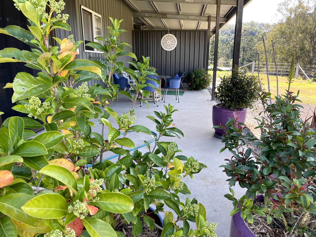 A covered outdoor area is surrounded by potted plants and greenery. A circular wall decoration is visible on the wall of the cottage. Several chairs are arranged on the porch, creating a serene spot overlooking the landscape.
