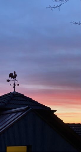 A weather vane featuring a rooster is positioned atop a roof, silhouetted against a colorful sky at dusk. Hues of purple, pink, and orange blend seamlessly, creating a serene backdrop for the structure. The sloped roof adds depth to the scene.