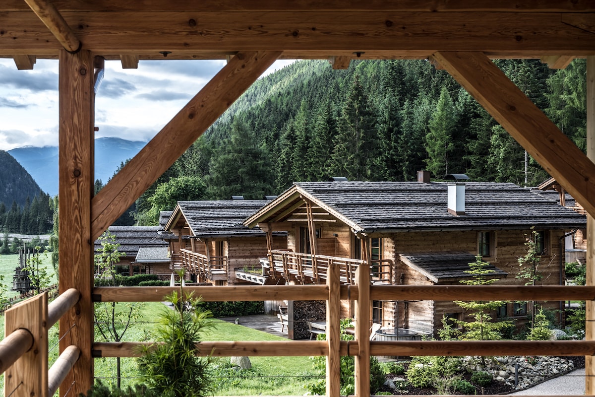 A view from a balcony showcases rustic wooden chalets nestled among lush greenery. Natural light illuminates the surroundings, while the mountains rise in the background, enhancing the peaceful atmosphere. The wooden railing frames the picturesque scene, inviting relaxation in a tranquil setting.