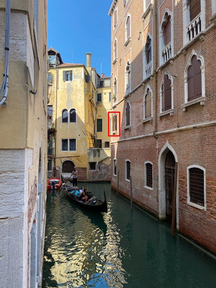 A narrow Venetian canal is visible, flanked by historic buildings featuring typical architecture. A gondola floats quietly along the water, with passengers enjoying the view. Sunlight reflects off the water's surface, creating a serene atmosphere amidst the charming surroundings.