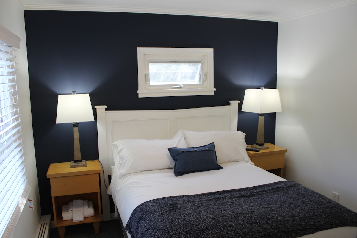 A cozy room featuring a full bed adorned with white linens and a navy throw blanket. Bedside tables with lamps provide soft lighting beside the bed, while a small window allows natural light to filter in. Neatly stacked towels can be seen below the tables.