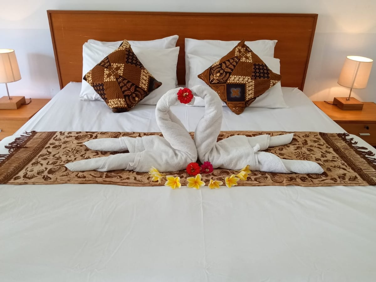A spacious bed is adorned with elegant towel art shaped like swans, surrounded by fresh flowers. Decorative pillows with intricate patterns are placed at the head of the bed, complemented by side lamps providing soft lighting.