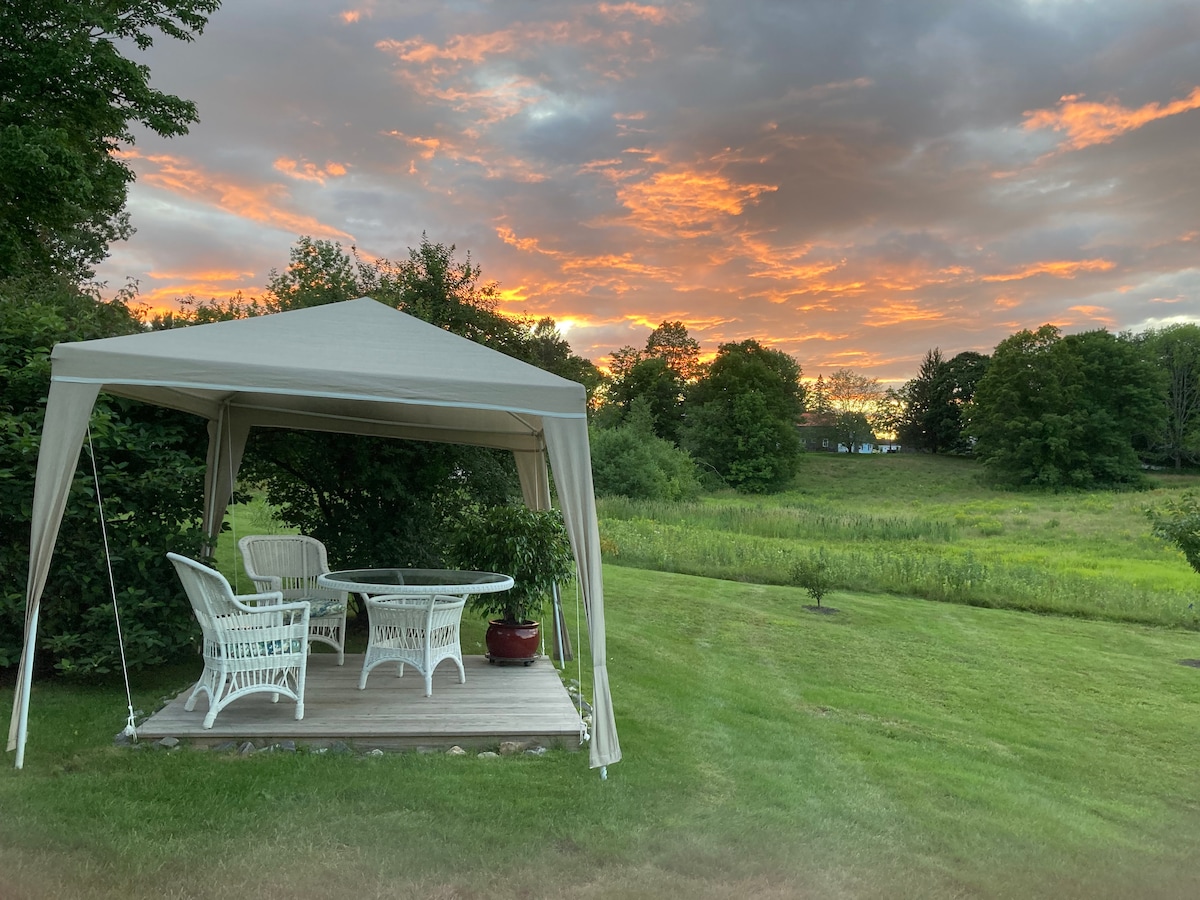 A shaded patio area features a white gazebo with a wicker table and chairs set on a wooden deck. Behind, a picturesque landscape showcases lush green foliage and an expansive field, all under a vibrant sunset sky filled with soft orange and pink hues.