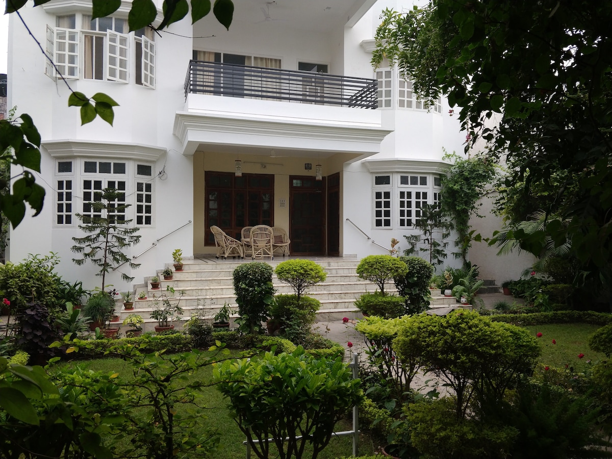 A heritage villa is presented with a spacious, welcoming staircase leading to the entrance. Lush greenery and flowering plants surround the steps, while a cozy seating area with wicker chairs invites relaxation. Large windows enhance the villa's bright and airy ambiance.