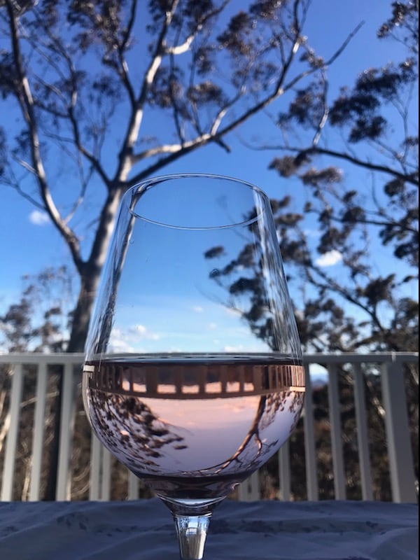A clear wine glass filled with rose reflects the trees and sky. The background features a blue sky scattered with clouds and tall trees, creating a serene outdoor setting. The glass stands on a table, enhancing the connection between nature and relaxation.